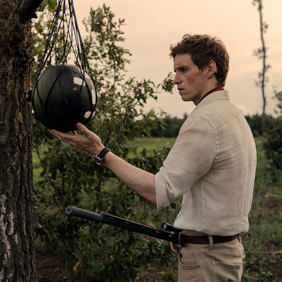 In a scene from The Day of the Jackal, a smartly dressed man (Eddie Redmayne) stand by a tree holding a rifle. 